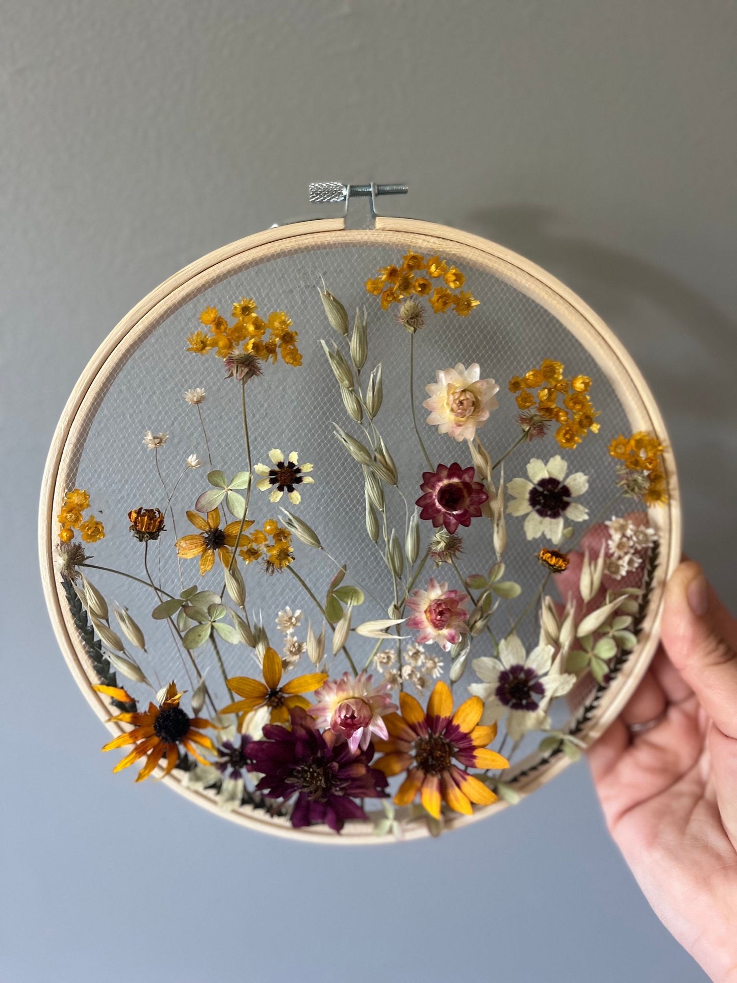 Real dried flowers are embroidered on tulle secured by an embroidery hoop. Dried botanicals include zinnias, starflowers, strawflowers and hydrangeas. The hoop is hung from the wall as a piece of wall art. Hoop is 8 inches in diameter.