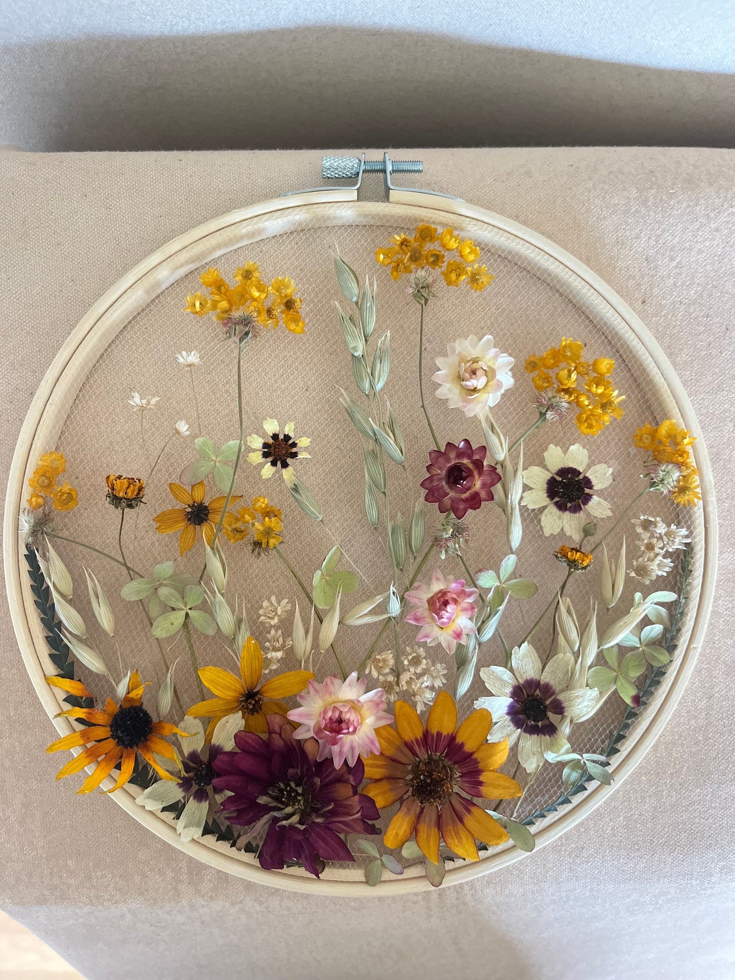 Real dried flowers are embroidered on tulle secured by an embroidery hoop. Dried botanicals include zinnias, starflowers, strawflowers and hydrangeas. The hoop is hung from the wall as a piece of wall art. Hoop is 8 inches in diameter.