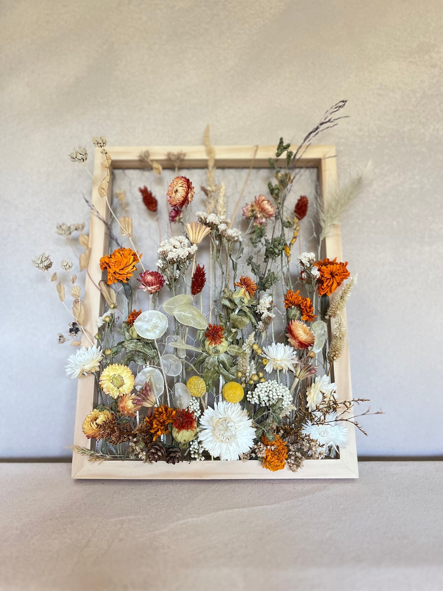 Fall dried flowers hang from a wooden frame secured by metal netting. Dried botanicals include strawflowers, marigolds, Zanzibar, lunaria and grasses. The frame is hung from the wall as a piece of wall art. Frame measures 11x14 and approx 5/8 deep.