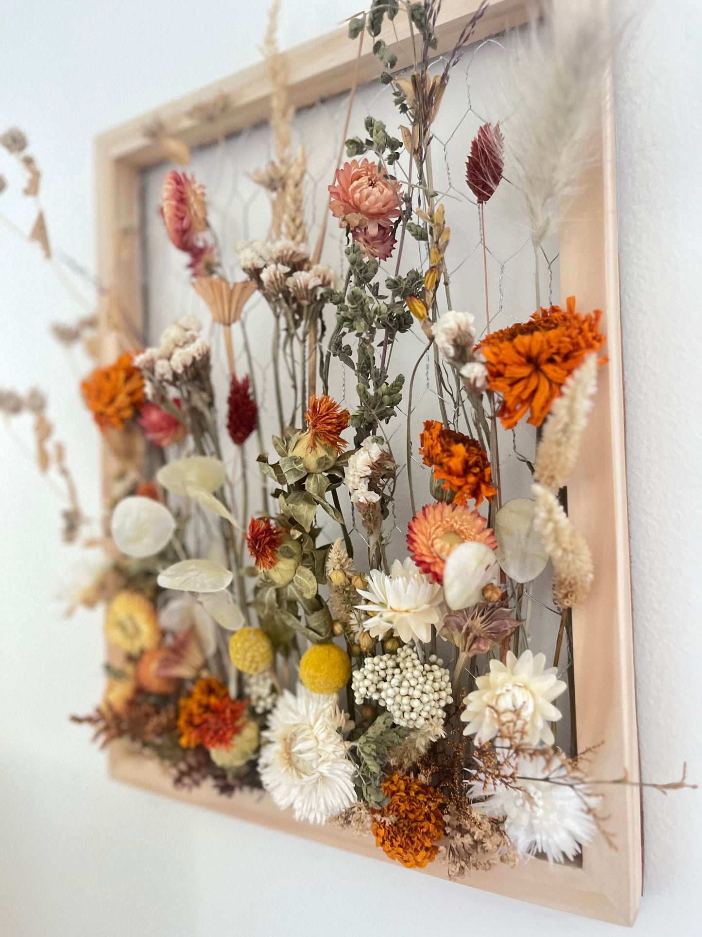 Fall dried flowers hang from a wooden frame secured by metal netting. Dried botanicals include strawflowers, marigolds, Zanzibar, lunaria and grasses. The frame is hung from the wall as a piece of wall art. Frame measures 11x14 and approx 5/8 deep.