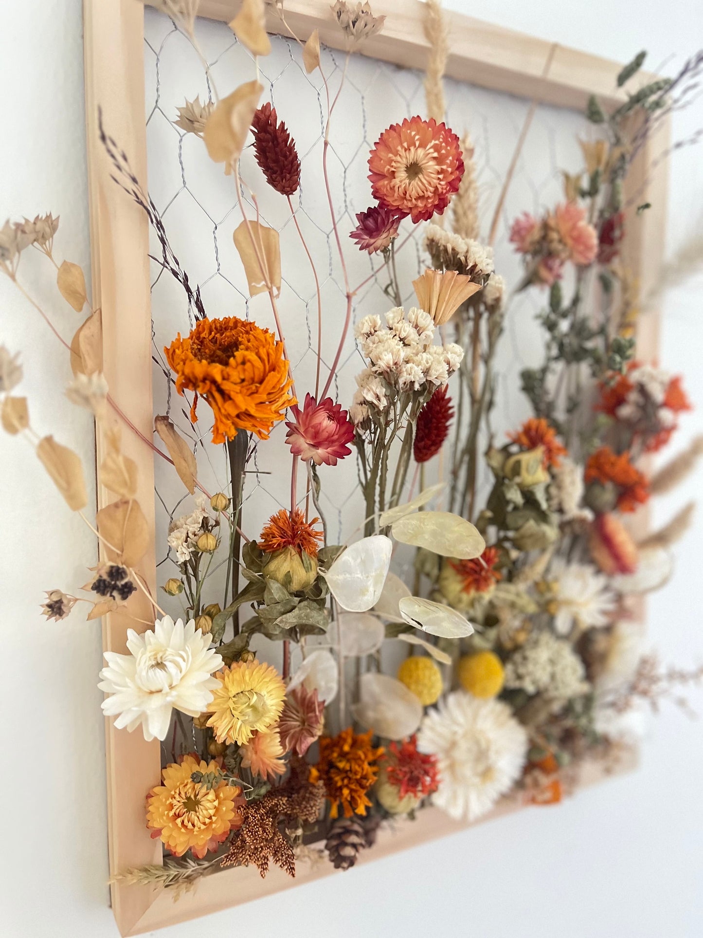 Fall dried flowers hang from a wooden frame secured by metal netting. Dried botanicals include strawflowers, marigolds, Zanzibar, lunaria and grasses. The frame is hung from the wall as a piece of wall art. Frame measures 11x14 and approx 5/8 deep.