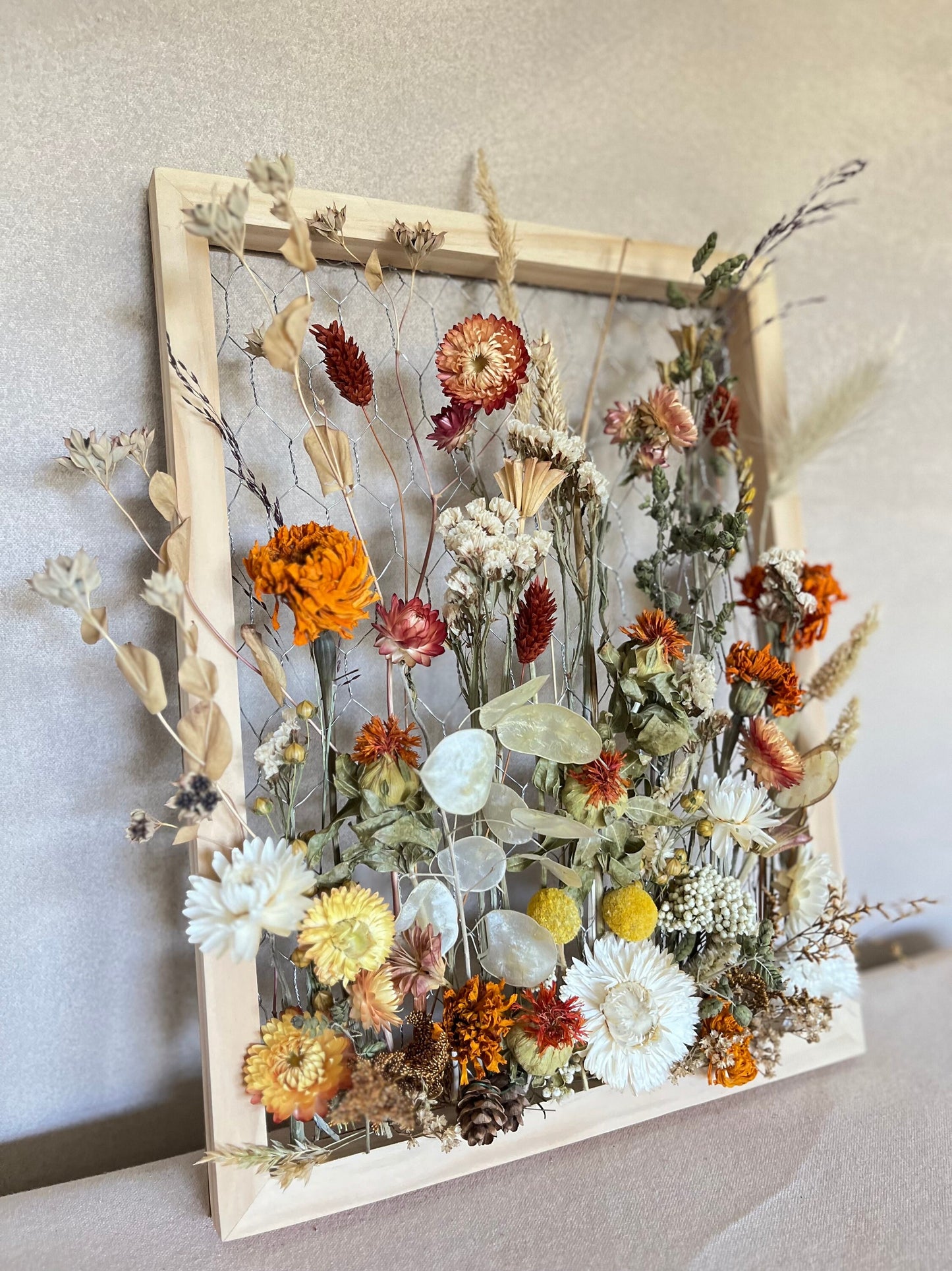 Fall dried flowers hang from a wooden frame secured by metal netting. Dried botanicals include strawflowers, marigolds, Zanzibar, lunaria and grasses. The frame is hung from the wall as a piece of wall art. Frame measures 11x14 and approx 5/8 deep.