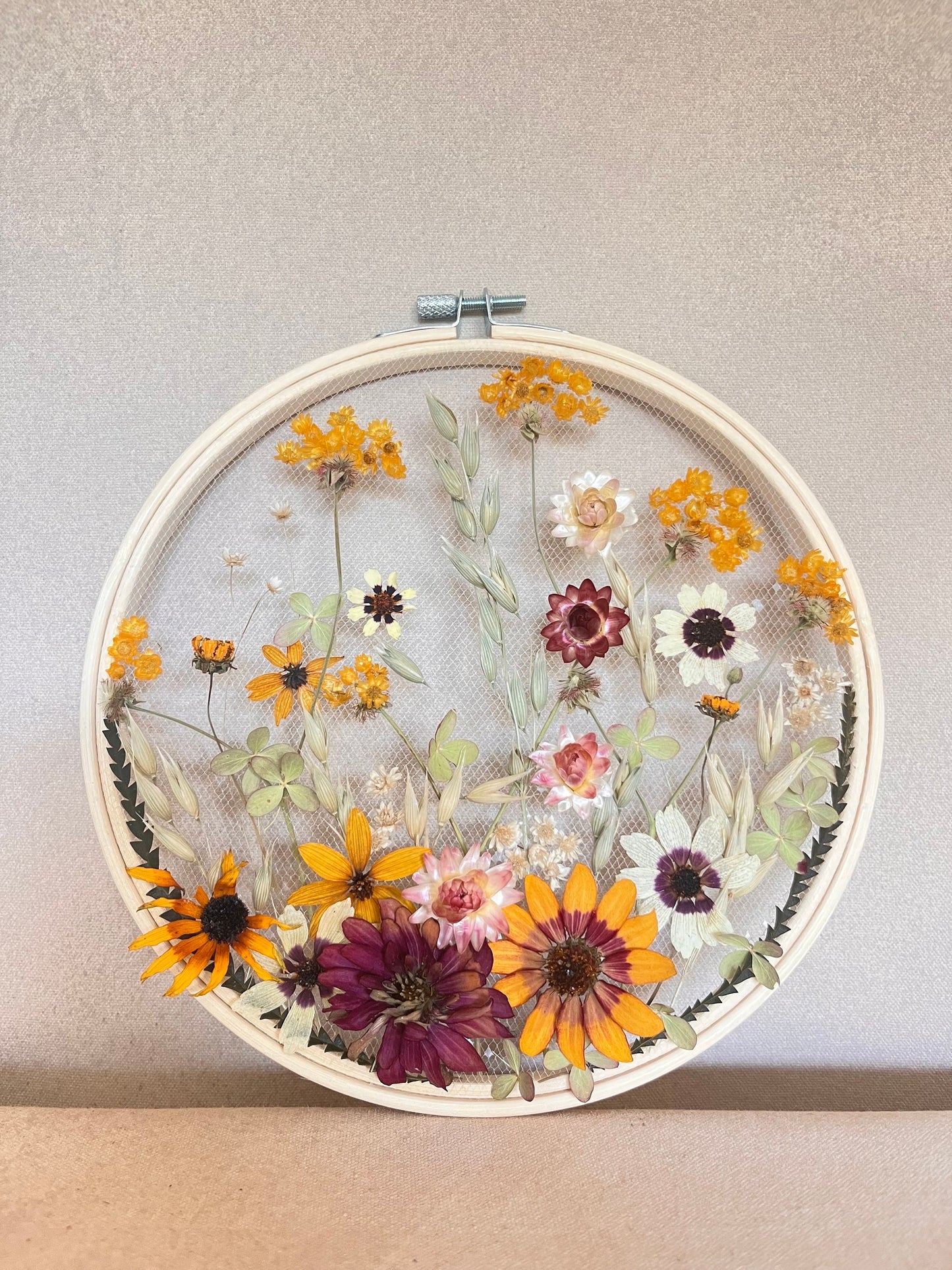 Real dried flowers are embroidered on tulle secured by an embroidery hoop. Dried botanicals include zinnias, starflowers, strawflowers and hydrangeas. The hoop is hung from the wall as a piece of wall art. Hoop is 8 inches in diameter.