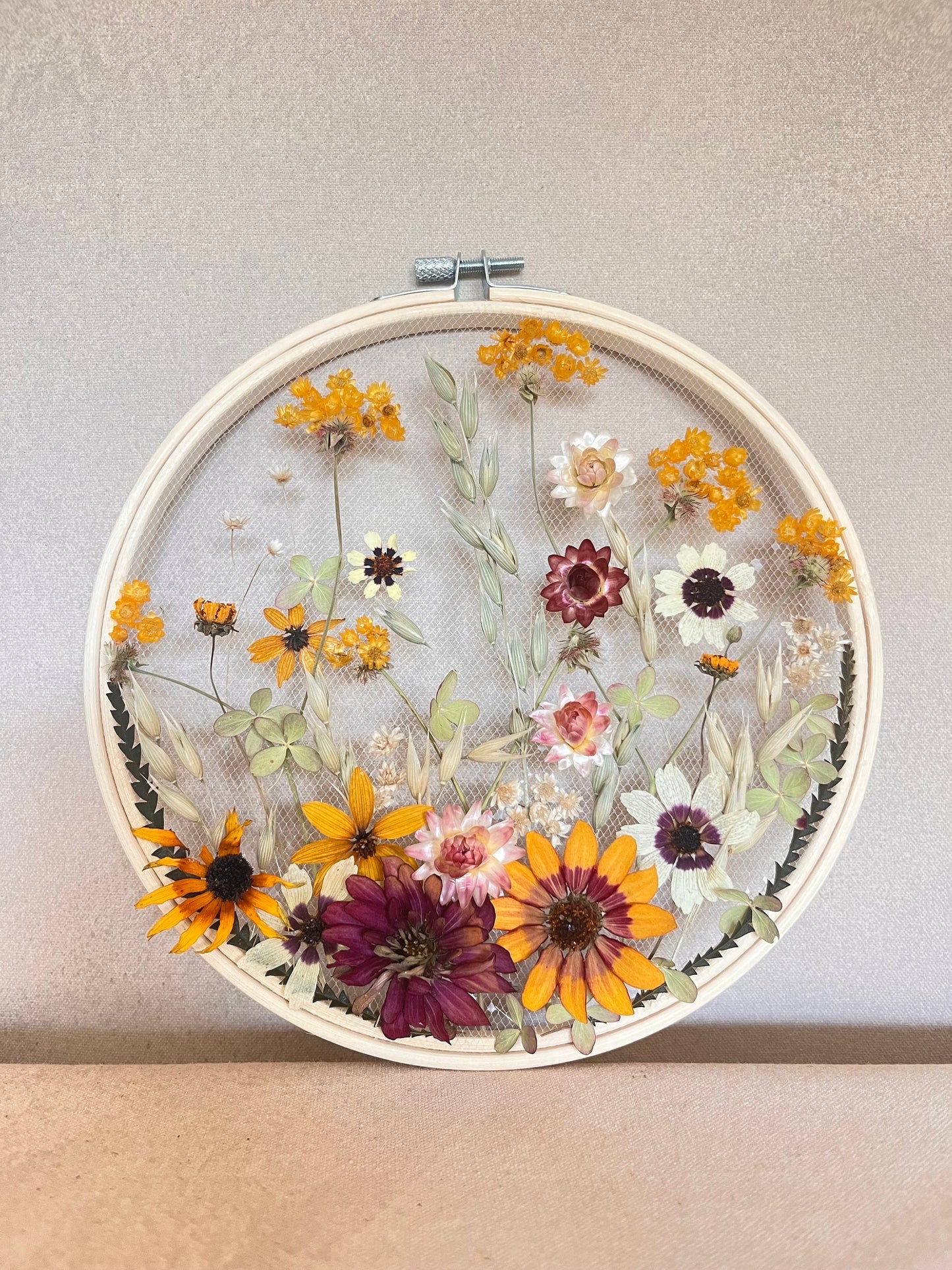 Real dried flowers are embroidered on tulle secured by an embroidery hoop. Dried botanicals include zinnias, starflowers, strawflowers and hydrangeas. The hoop is hung from the wall as a piece of wall art. Hoop is 8 inches in diameter.