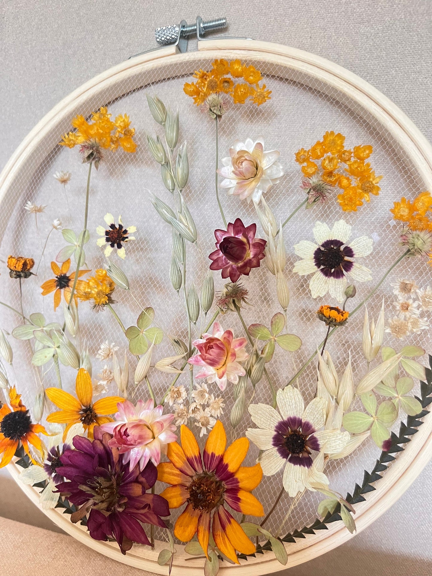 Real dried flowers are embroidered on tulle secured by an embroidery hoop. Dried botanicals include zinnias, starflowers, strawflowers and hydrangeas. The hoop is hung from the wall as a piece of wall art. Hoop is 8 inches in diameter.