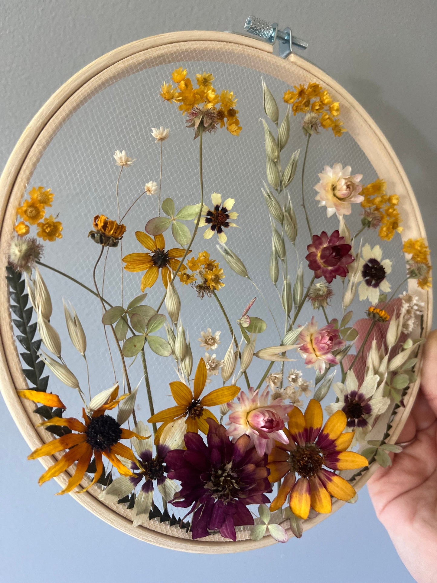Real dried flowers are embroidered on tulle secured by an embroidery hoop. Dried botanicals include zinnias, starflowers, strawflowers and hydrangeas. The hoop is hung from the wall as a piece of wall art. Hoop is 8 inches in diameter.