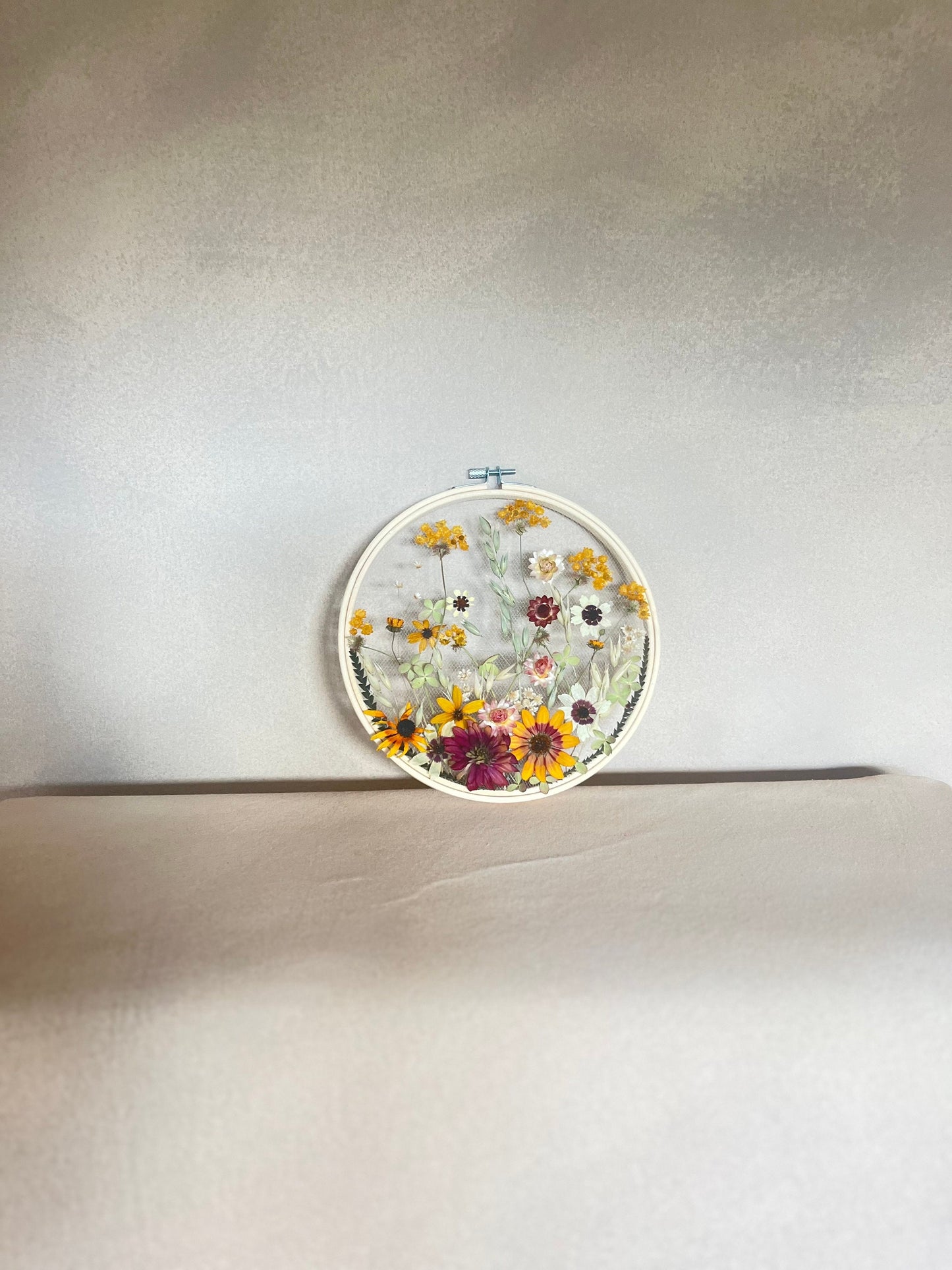 Real dried flowers are embroidered on tulle secured by an embroidery hoop. Dried botanicals include zinnias, starflowers, strawflowers and hydrangeas. The hoop is hung from the wall as a piece of wall art. Hoop is 8 inches in diameter.