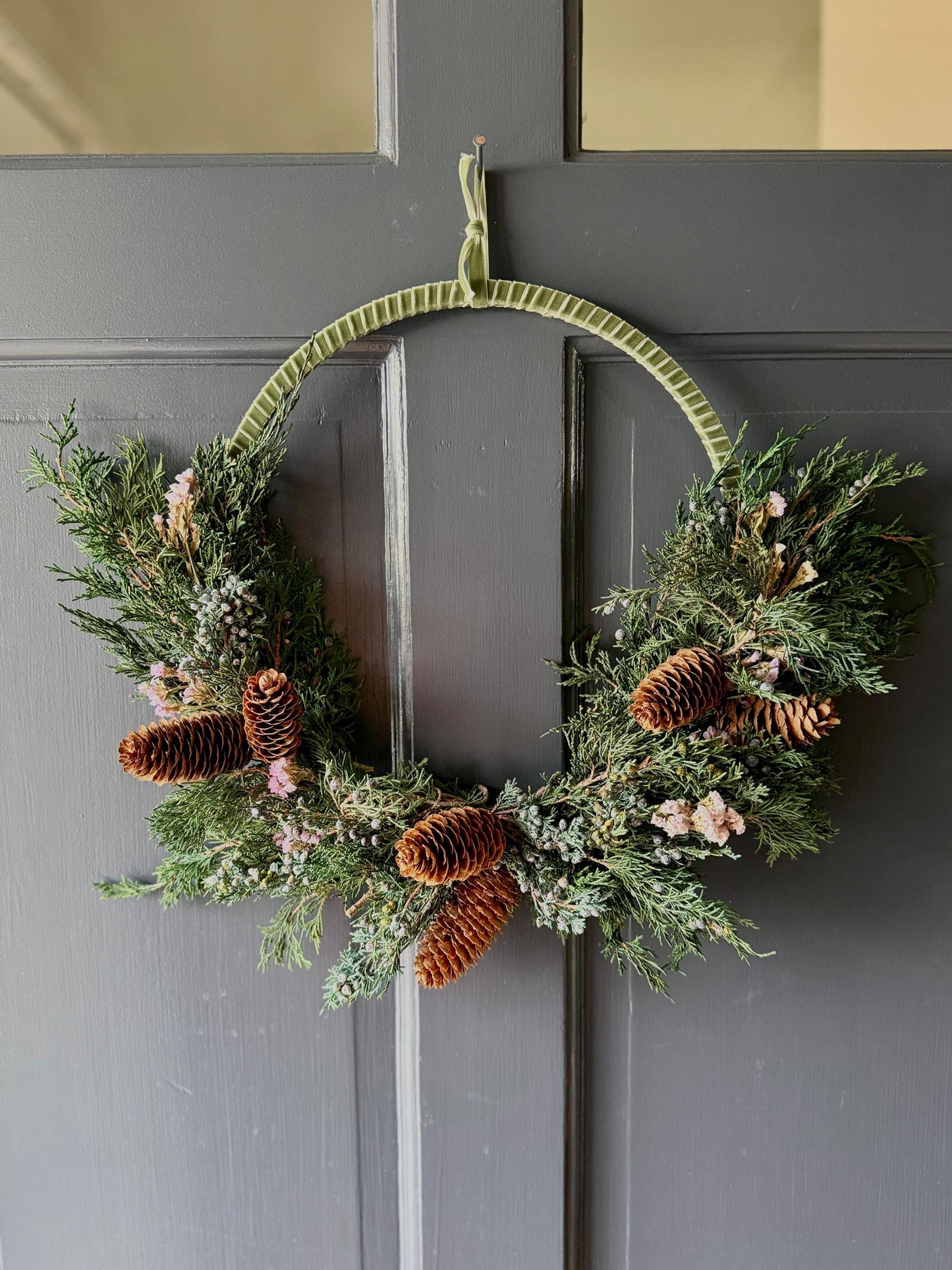 Holiday wreath featuring preserved juniper, lavender statice, pinecones and a beautiful pine green velvet ribbon. Adorn a wall, mantel or sheltered door; this dried flower wreath is the perfect addition to your holiday decor.