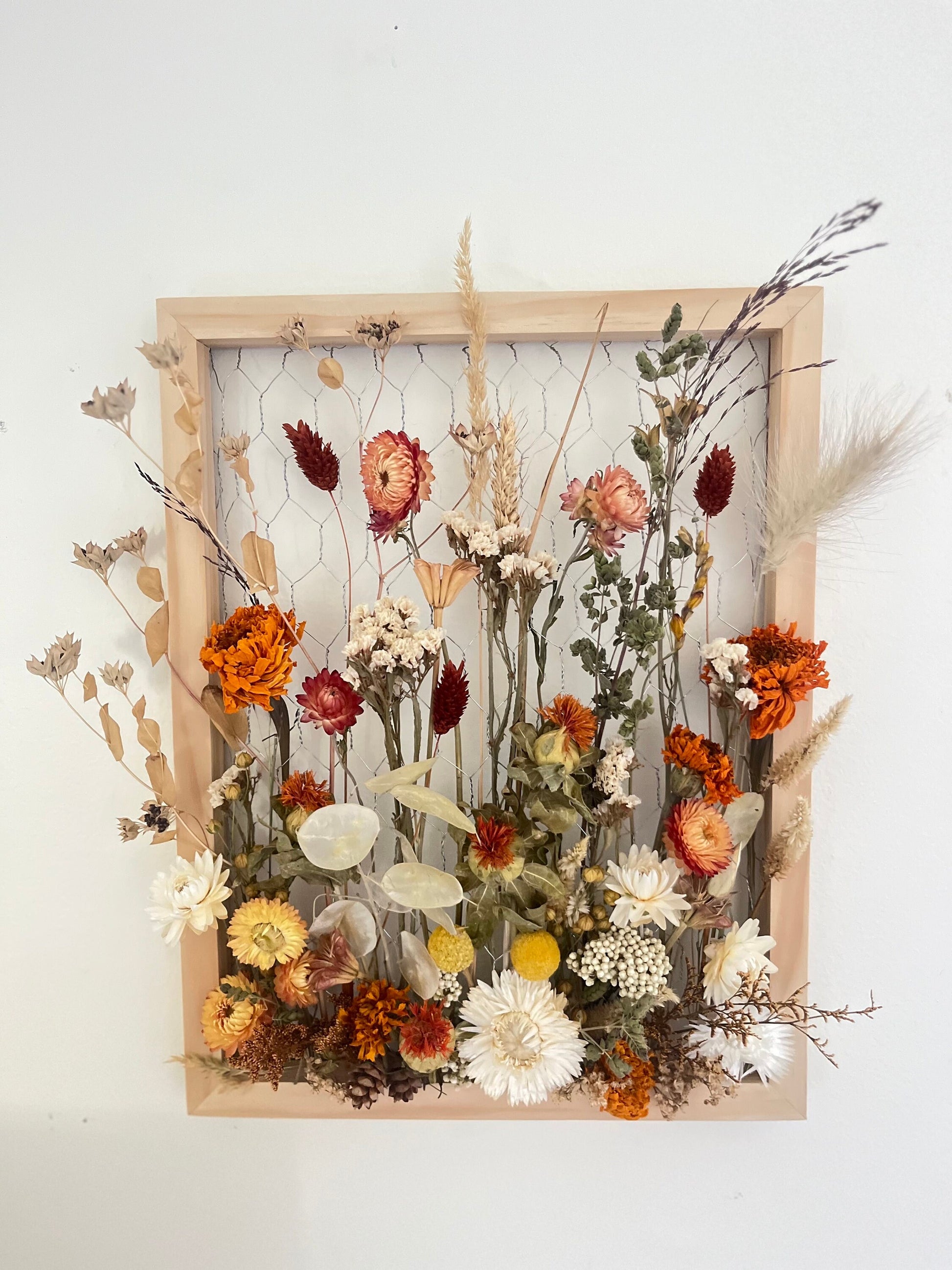 Fall dried flowers hang from a wooden frame secured by metal netting. Dried botanicals include strawflowers, marigolds, Zanzibar, lunaria and grasses. The frame is hung from the wall as a piece of wall art. Frame measures 11x14 and approx 5/8 deep.