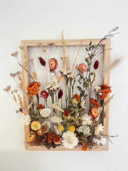 Fall dried flowers hang from a wooden frame secured by metal netting. Dried botanicals include strawflowers, marigolds, Zanzibar, lunaria and grasses. The frame is hung from the wall as a piece of wall art. Frame measures 11x14 and approx 5/8 deep.