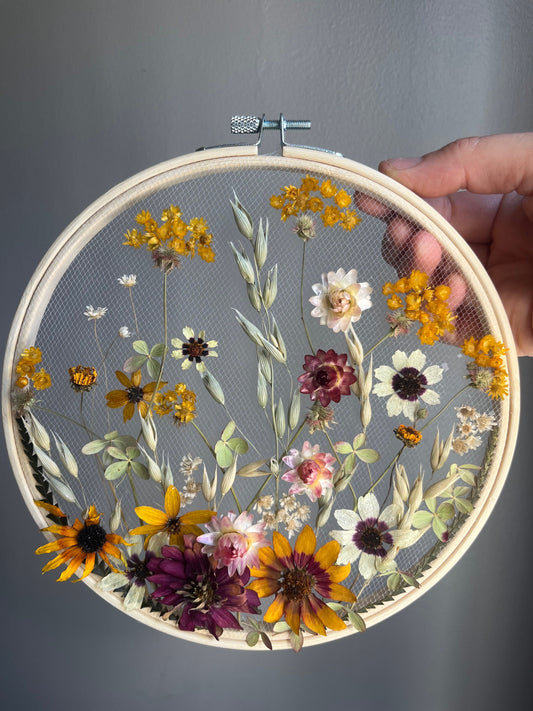 Real dried flowers are embroidered on tulle secured by an embroidery hoop. Dried botanicals include zinnias, starflowers, strawflowers and hydrangeas. The hoop is hung from the wall as a piece of wall art. Hoop is 8 inches in diameter.