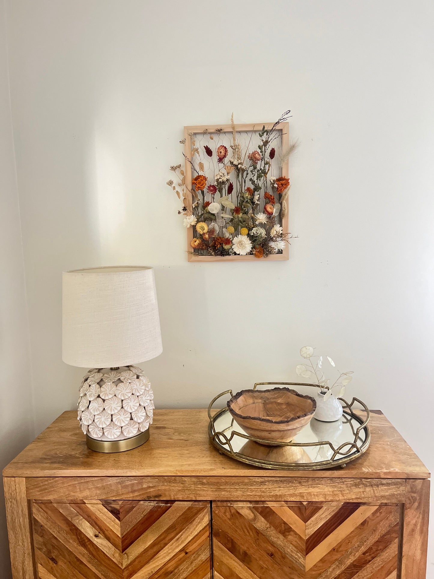 Fall dried flowers hang from a wooden frame secured by metal netting. Dried botanicals include strawflowers, marigolds, Zanzibar, lunaria and grasses. The frame is hung from the wall as a piece of wall art. Frame measures 11x14 and approx 5/8 deep.