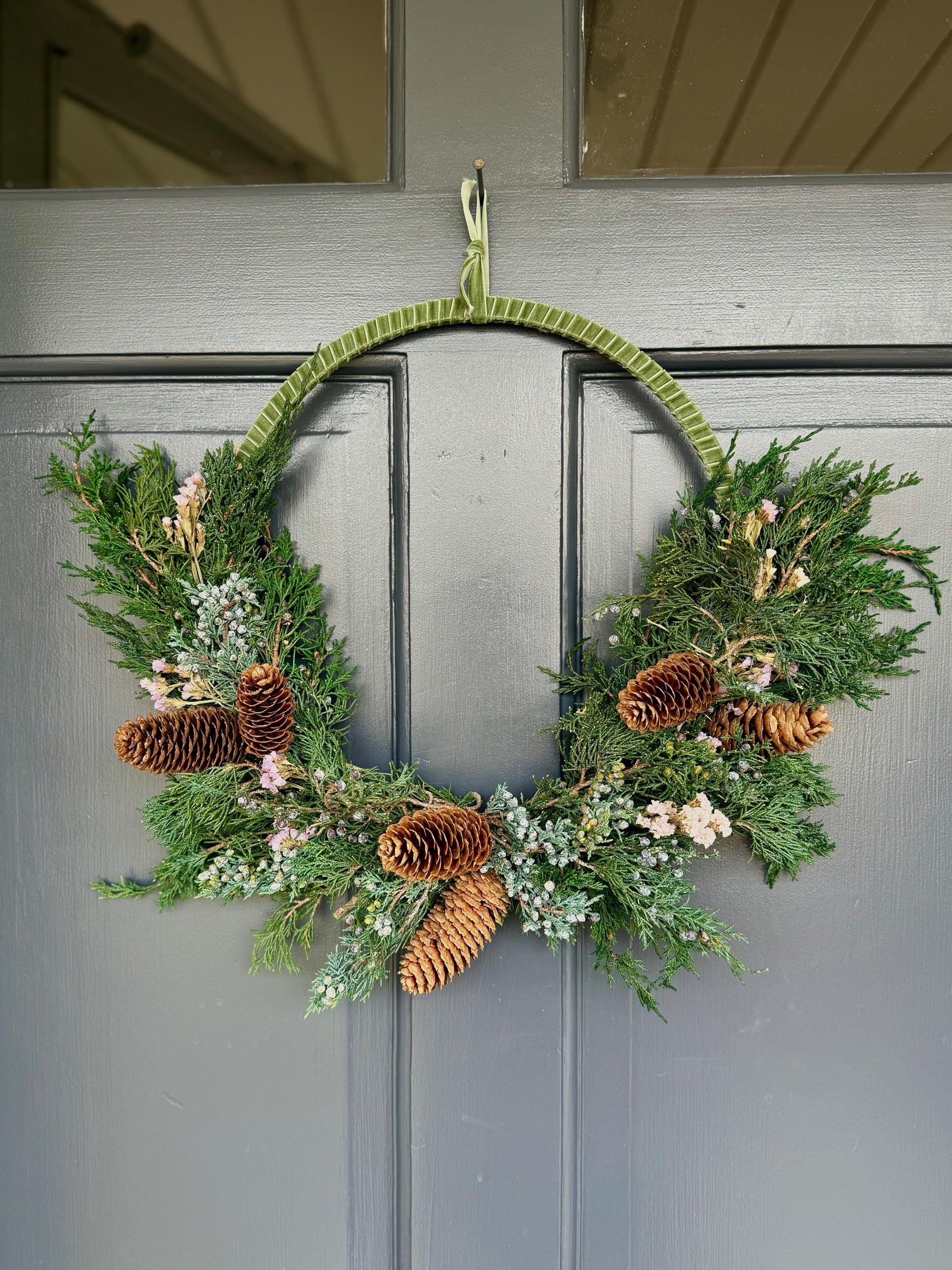 Holiday wreath featuring preserved juniper, lavender statice, pinecones and a beautiful pine green velvet ribbon. Adorn a wall, mantel or sheltered door; this dried flower wreath is the perfect addition to your holiday decor.