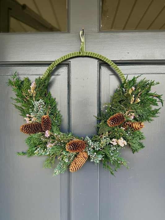 Holiday wreath featuring preserved juniper, lavender statice, pinecones and a beautiful pine green velvet ribbon. Adorn a wall, mantel or sheltered door; this dried flower wreath is the perfect addition to your holiday decor.